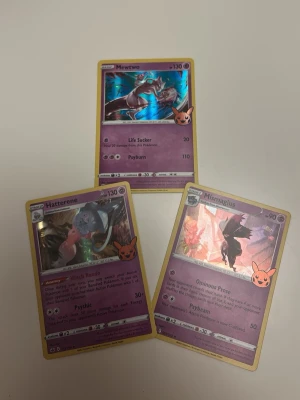 Pokémonkort Mewtwo Hatterene Mismagius, Hollow - Tre Pokémonkort: Mewtwo, Hatterene och Mismagius. Alla korten har lila bakgrund och guldkant, samt en Pikachu-stämpel i hörnet. Motiven är färgstarka och detaljerade, med hollow effekter på vissa delar. 