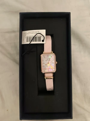 Rosa klocka från Daniel Wellington  - Stilren klocka från Daniel Wellington med fyrkantig urtavla i pärlemor och guldfärgade detaljer. Klockan har ett ljusrosa tygarmband och elegant design som passar perfekt till din outfit.