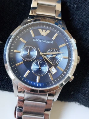 Emporio Armani blå klocka i stål - Snygg klocka från Emporio Armani med blå urtavla och silverfärgat armband i rostfritt stål. Klockan har tre mindre urtavlor, datumvisning och klassisk design med märkets logga högst upp. Perfekt accessoar för en stilren look.