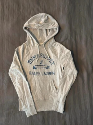 Grå hoodie från Ralph Lauren - Snygg grå hoodie från Ralph Lauren med blått tryck på bröstet där det står 'DNM & SUPPLY' och 'RALPH LAUREN'. Hoodien har huva med dragsko och en klassisk känguruficka framtill. Perfekt för en avslappnad och cool stil. Längd 57cm bredd 40cm. Det är Xs men passar Xxs.