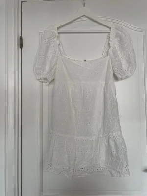 Vit kort klänning med puffärm - Superfin vit kort klänning med broderade blommor och puffärmar. Klänningen har en söt volang nertill. Perfekt för student!