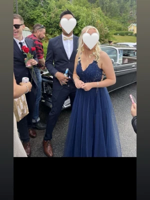 Marinblå långklänning med spets - Elegant marinblå långklänning med spetsdetaljer upptill och v-ringad hals. Klänningen har tunna axelband och ett flödande, transparent tyg i kjolen som ger ett lyxigt intryck. Perfekt för bal eller student. Materialet känns som polyester och spets.