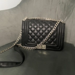 Svart handväska - Snygg svart axelväska med quiltad framsida och silverfärgad kedja som axelrem. Väskan har en rektangulär form och ett stilrent metallspänne framtill. Perfekt för dig som gillar klassisk och trendig design. Tror att den är från ur & penn 