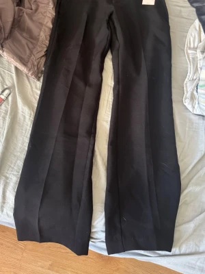 Svarta vida kostymbyxor - Snygga svarta kostymbyxor med vida ben och pressveck. Byxorna har diskreta bakfickor och hällor för bälte. Perfekta för dig som vill ha en stilren och klassisk look. Materialet är slätt och känns bekvämt mot huden.