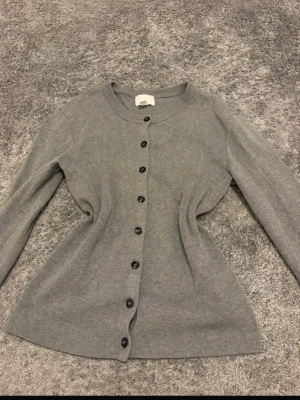 Grå basic kofta med knappar - En enkel och stilren grå kofta med rund halsringning och svarta knappar hela vägen framtill. Koftan har lång ärm och är tillverkad i ett mjukt, tunt stickat material som passar perfekt till lager på lager. Klassisk modell som funkar till det mesta.