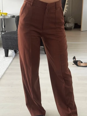 Bruna vida kostymbyxor - Snygga bruna kostymbyxor med hög midja och raka, vida ben. Byxorna har pressveck framtill, bälteshällor och diskreta sidofickor. Perfekta för en trendig och stilren look. Materialet ser ut att vara mjukt och följsamt, troligen bomullsblandning. Short 