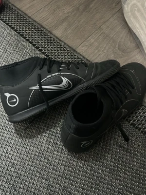Svarta Nike Mercurial fotbollsskor - Säljer ett par svarta Nike Mercurial fotbollsskor med vita detaljer och logga. Skorna har högre skaft för extra stöd, snörning och slitstark yttersula för bra grepp på planen. Snygg design med grafiska linjer och texten 'Engineered for speed'. Perfekta för dig som vill ha stil och funktion på planen. Helt nytt har använts Max 6 ggr jätte bra skick