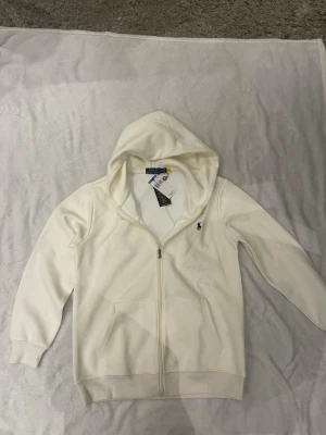 Vit hoodie från Polo Ralph Lauren - Snygg vit hoodie från Polo Ralph Lauren med dragkedja framtill och klassisk huva. Tröjan har två fickor fram och den ikoniska lilla marinblå loggan broderad på bröstet. Tillverkad i mjukt bomullsmaterial som känns skönt mot huden. Perfekt för en clean och stilren look.