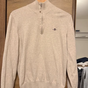 Beige halvzip-tröja från Gant - Snygg beige stickad tröja från Gant med halv dragkedja och broderad logga på bröstet. Tröjan har hög krage och ribbade muddar vid ärmslut och nederkant. Perfekt för en stilren och avslappnad look.