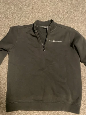 Svart Sail Racing tröja half zip - Svart långärmad tröja från Sail Racing med half zip och hög krage. Tröjan har en stilren logga på bröstet och är tillverkad i mjukt material, perfekt för en avslappnad och sportig look. Pefekt till våren! Passar folk mellan 154-160cm