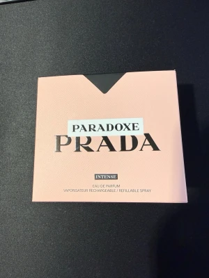 Prada Paradoxe Intense Eau de Parfum - Prada Paradoxe Intense Eau de Parfum kommer i en snygg trekantig glasflaska med svart lock och rosa-orange doftvätska. Förpackningen är ljusrosa med svart och silverdetaljer. En lyxig och modern parfym som sticker ut i badrumshyllan.