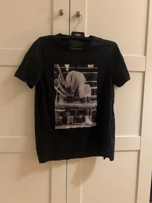 Svart t-shirt med boxningsmotiv Boss - Svart t-shirt från limitato med ett stort tryck framtill i sammet material som visar mohammed ali i ringen. Klassisk passform och rund hals. Tillverkad i mjuk bomull som känns skön mot huden. Perfekt för dig som gillar sportiga och stilrena plagg.