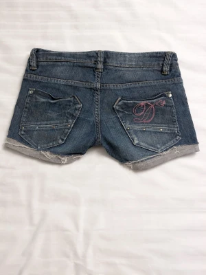 Blå jeansshorts med cool design  - Säljer ett par blå jeansshorts från Detroit med råa kanter och slitna detaljer. Shortsen har fem fickor, silverfärgade nitar och en rosa dragkedja fram. På ena bakfickan finns ett broderat D i rosa. Perfekta för sommaren!