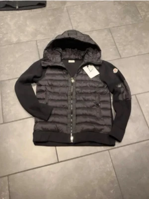 Svart cardigan från Moncler - Snygg svart cardigan från Moncler med huva och dragkedja framtill. Cardigan har quiltad front i glansigt material och stickade ärmar samt ribbade muddar. Klassisk Moncler-logga på ärmen och praktiska sidofickor med dragkedja. Perfekt för kalla vinterdagar.