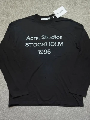  Acne Studios - Svart långärmad t-shirt från Acne Studios med tryck i vitt framtill där det står 'Acne Studios STOCKHOLM 1996'. Klassisk rund hals och normal passform. Perfekt för dig som gillar stilrena och trendiga plagg.