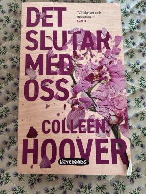 Det slutar med oss av Colleen Hoover - En gripande roman av Colleen Hoover med ett omslag i ljus träfärg och lila text samt blomblad. Boken är på svenska och handlar om Lily, kärlek och svåra val. Pocketutgåva från Lovereads, perfekt för dig som gillar starka berättelser och känslor.