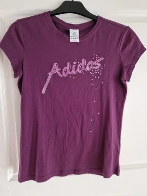 Lila Adidas t-shirt med strassdetaljer - Snygg lila t-shirt från Adidas med korta ärmar och coolt strassmönster i rosa på bröstet. T-shirten har en klassisk passform och är gjord i mjuk bomull, perfekt för en avslappnad och sportig stil.