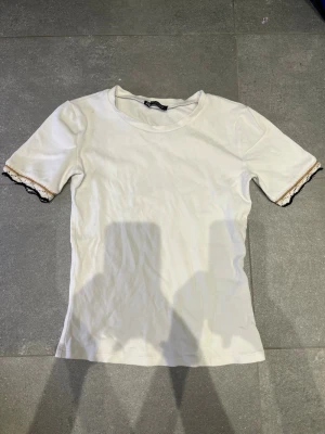 Vit t-shirt med spetsdetaljer - Snygg vit t-shirt med rund hals och korta ärmar. Ärmsluten har unika spetsdetaljer i svart och beige som ger en cool kontrast. T-shirten har en rak passform och är tillverkad i mjukt material, perfekt för en avslappnad stil. STORLEK S MEN PASSAR ÄVEN XS