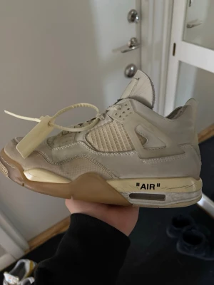 Off-White x Air Jordan 4 Sail - Säljer ett par Off-White x Air Jordan 4 Sail sneakers i beige och krämvitt. Skorna har meshpaneler, genomskinliga detaljer och den ikoniska 'AIR'-texten på mellansulan. De har en chunky sula och Off-White zip-tie. Perfekta för dig som gillar streetwear och unika samarbeten.