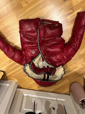 Röd dunjacka från Moncler - Snygg röd dunjacka från Moncler med glansig finish och beige insida. Jackan har huva, dragkedja, tryckknappar och flera fickor. Insidan har randiga detaljer i rött, vitt och blått. Perfekt för kalla dagar och riktigt trendig look.sjölvklart är den äkta