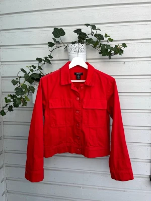 Röd jeansjacka från Lindex - Snygg röd jeansjacka från Lindex med klassisk krage, två bröstfickor med lock och knappar framtill. Jackan har en rak, något kortare passform och långa ärmar. Perfekt för dig som vill sticka ut med färg i din outfit.