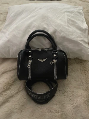 Svart Zadig & Voltaire handväska - Cool svart handväska i läder från Zadig & Voltaire med silverfärgade dragkedjor, dubbla handtag och bred axelrem med VOLTAIRE-tryck. Väskan har en snygg logga framtill och detaljerade nitar längs kanterna. Perfekt för dig som gillar edgy och trendig stil.