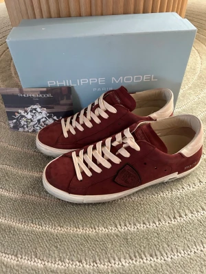 Vinröda Philippe Model sneakers - Säljer ett par snygga vinröda Philippe Model sneakers i mocka med vita detaljer och vita skosnören. Skorna har en låg profil, vit sula och beige hälparti. Klassisk Philippe Model-logga på sidan och bekväm innersula. Perfekta för dig som vill ha stilrena och trendiga sneakers.