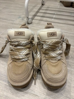 Beige fodrade sneakers från UGG - Snygga beige sneakers från UGG med breda, mönstrade snören och fluffigt foder i fuskpäls. Ovandelen är i mocka och mesh, vilket ger en cool och sportig look. Perfekta för dig som vill ha både stil och komfort. Skorna är endast använda en gång. Obs! Ett fäste till ena skons skosnöre är trasigt. Därav det låga priset! Se sista bilden. 