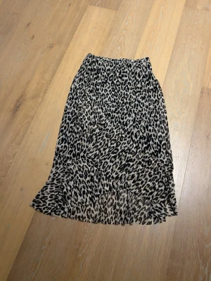 Leopardmönstrad kjol från Lindex - Snygg leopardmönstrad kjol från Lindex med volang nertill. Kjolen har en beige och svart färgskala och är tillverkad i ett lätt och luftigt material. Perfekt för dig som vill sticka ut med ett trendigt djurmönster.
