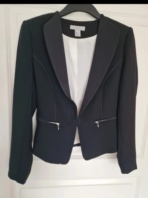 Svart kavaj med dragkedjor - Stilren svart kavaj med två coola dragkedjor framtill. Kavajen har figurnära passform och är fodrad med vitt tyg.