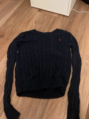 Mörkblå kabelstickad tröja Polo Ralph Lauren - Kabelstickad mörkblå tröja från Polo Ralph Lauren i mjuk pima cotton. Tröjan har rund halsringning, långa ärmar och den klassiska röda loggan broderad på bröstet. Perfekt för en stilren och tidlös look.