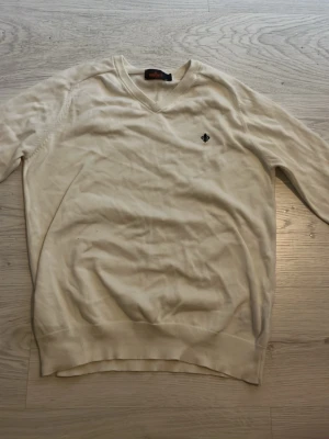 Beige stickad tröja från Morris - Snygg beige stickad tröja från Morris med V-ringning och diskret logga broderad på bröstet. Tröjan har långa ärmar och ribbade muddar vid ärmslut och nederkant. Perfekt för en stilren och enkel look.