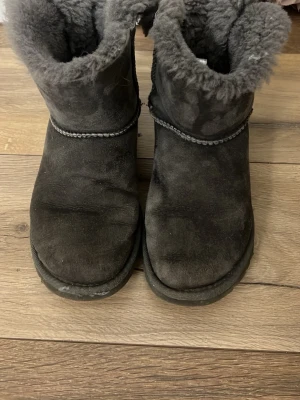 Grå UGG Australia fårskinnsstövlar - Mysiga grå stövlar från UGG Australia i äkta fårskinn med fluffigt foder och dekorativa rosetter bak. Klassisk rund tå och mellanhögt skaft. Perfekta för kalla dagar när du vill hålla fötterna varma och snygga.