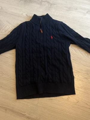 Mörkblå kabelstickad tröja Polo Ralph Lauren - Mörkblå kabelstickad tröja från Polo Ralph Lauren med halv dragkedja och hög krage. Tröjan har det klassiska röda Polo-logot på bröstet och ribbade muddar vid ärmslut och nederkant. Perfekt för en stilren och avslappnad look.