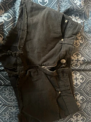 Svarta jeansshorts med rå kant - Säljer ett par svarta jeansshorts med fransig, rå kant och klassisk femficksdesign. Shortsen har dragkedja och knapp framtill samt bälteshällor. Perfekta för sommaren och ger en avslappnad vibe.