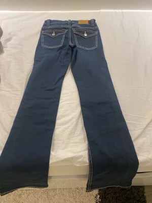 Blå bootcut jeans från Gina Tricot - Säljer ett par mörkblå bootcut jeans från Gina Tricot med kontrasterande vita sömmar och snygga fickdetaljer bak. Jeansen har breda benslut och klassisk femficksdesign. Perfekta för dig som gillar en retroinspirerad look.