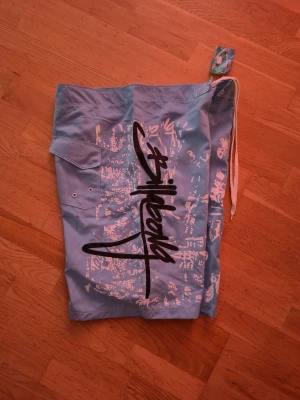 Blå Billabong badshorts med tryck - Säljer ett par blå badshorts från Billabong med coolt grafiskt tryck i vitt, svart och rött. Shortsen har snörning i midjan, en ficka med lock och ventilationshål på sidan. Materialet är lätt polyester, perfekt för strand eller pool.