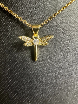 Guldigt halsband med trollslända - Snyggt halsband med tunn kedja i guldfärg och ett hängsmycke formad som en trollslända. Vingarna är dekorerade med små glittrande stenar som ger extra detalj. Perfekt för dig som gillar unika accessoarer med naturtema.