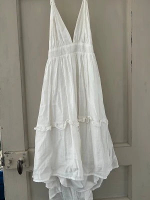 Vit omlottklänning med volang - Fin klänning i omlottmodell med v-ringning och volangdetaljer. Klänningen har smala axelband som knyts i nacken och är tillverkad i ett lätt tyg, perfekt för varma dagar. Passformen är loose nertill men kan justeras på 3 olika ställen midja, rygg och nacke🙏🏻 2 lager så inte genomskinlig✨🥰