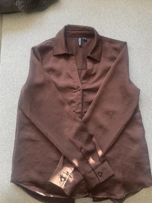 Brun skjorta från PIECES - Snygg brun skjorta från PIECES i klassisk modell med krage och knappar framtill. Skjortan har långa ärmar och en slits baktill😍
