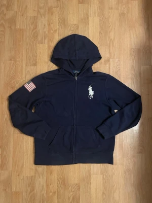 Marinblå zip hoodie Polo Ralph Lauren - Marinblå hoodie med dragkedja från Polo Ralph Lauren. Storlek 14-16 år, passar någon runt 160cm lång. Stor vit logga på bröstet, amerikansk flagga på ena ärmen och stor röd siffra 3 på ryggen. Klassisk huva och kängurufickor framtill. Tillverkad i mjukt bomullsmaterial.