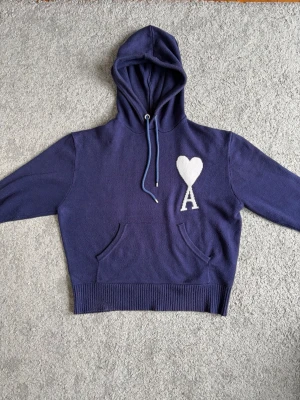 Mörkblå hoodie från Ami Paris - Snygg mörkblå hoodie från Ami Paris med vit hjärtlogga och bokstaven A på bröstet. Hoodien har dragsko i huvan, magficka och ribbade muddar i nederkant. Perfekt för en chill och trendig look.