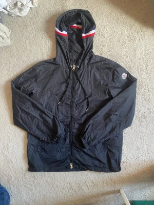 Svart vindjacka från Moncler - Snygg svart vindjacka från Moncler med huva och dragkedja framtill. Jackan har Moncler-logga på ärmen och röda samt vita detaljer på huvan. Tillverkad i lätt och tåligt material, perfekt för blåsiga dagar. Justerbar huva med snörning och elastiska muddar vid ärmslut. Storlek 3= M. Äkta
