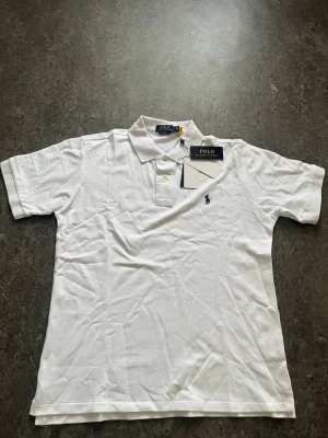 Polo piké - Klassisk vit polotopp från Polo Ralph Lauren med korta ärmar och broderad logga på bröstet. Tillverkad i mjuk bomull och har knappar vid kragen. Perfekt för en clean och stilren look.