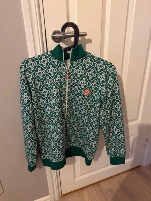 Grönmönstrad ziptröja från Casablanca  - Grön ziptröja från Casablanca med vitt grafiskt mönster över hela plagget. Tröjan har ribbade muddar, hög krage och vit dragkedja framtill. Liten röd logopatch på bröstet ger en extra detalj. Perfekt för dig som gillar retrostil och unika prints.
