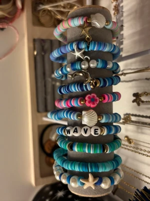Somriga pärlarmband med berlocker - Säljer ett set med elastiska armband i blå, turkosa, rosa och vita toner. Armbanden har platta pärlor, vissa med bokstäver, snäckor, stjärnor, delfin och blomma som berlocker. Perfekta för dig som gillar strandig och lekfull stil. Passar att mixa och matcha för en personlig look.