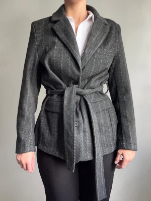 Grå kavaj med knytband - Stilren grå kavaj med fiskbensmönster och brett knytband i midjan. Klassisk krage, två framfickor och slits bak. Materialet ser ut att vara ullmix och ger en snygg, strukturerad look. Perfekt för dig som gillar tidlös och trendig stil.