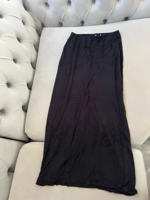 Svart långkjol från NA-KD - Säljer en svart långkjol från NA-KD i enkel och stilren design. Kjolen har en rak passform och är tillverkad i ett mjukt, tunt material som faller snyggt längs kroppen. Kjolen har en slits där bak så man kan röra på sig bekvämt.