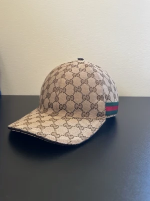 Beige GG-mönstrad keps från Gucci - Säljer en beige keps från Gucci med det klassiska GG-monogrammet över hela kepsen. På sidan finns den ikoniska gröna och röda randen. Kepsen har böjd skärm och är tillverkad i ett slitstarkt bomullsmaterial. Perfekt för dig som vill ha en stilren och lyxig accessoar. Använd en sommar.