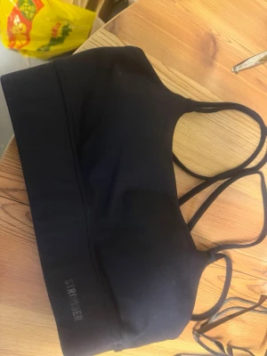 Svart sport-bh från Stronger - Snygg svart sport-bh från Stronger med dubbla smala axelband och bred resår under bysten. Tillverkad i stretchigt funktionsmaterial som passar perfekt för träning och aktivitet. Diskret logga på sidan av resåren. Använd två gånger 